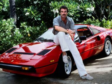 Tom Selleck sits on red car for a Magnum PI shoot. He's wearing white pants and a Hawaiian button-up shirt.
