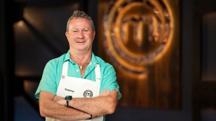 MasterChef Stephen Dennis crosses arms in front of MasterChef logo wearing white apron and blue collared shirt