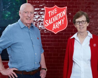 Ray Meagher and Salvos Chief Miriam Gluyas stand in front of red wall with The Salvation Army emblem between them
