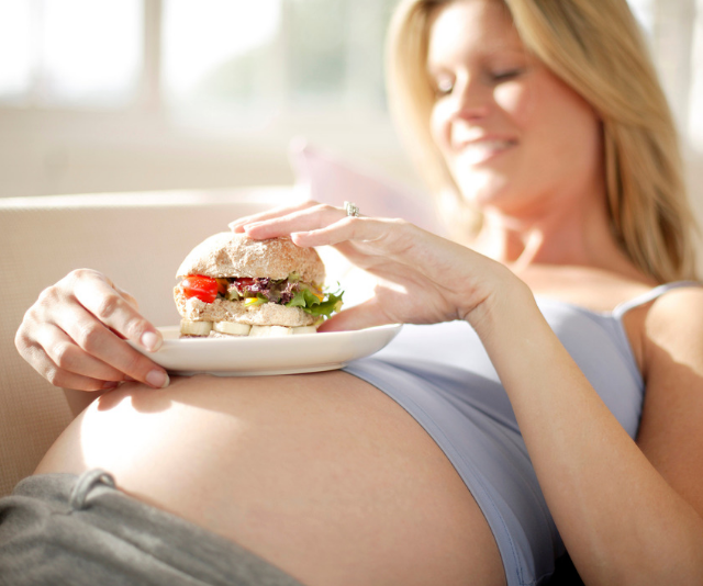 Pregnant woman laying down on lounge with salad sandwich resting on her baby bump.